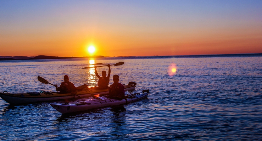 two kayaks rest in blue water while the sun sets behind them. one kayaker raises their paddle in the air in celebration.